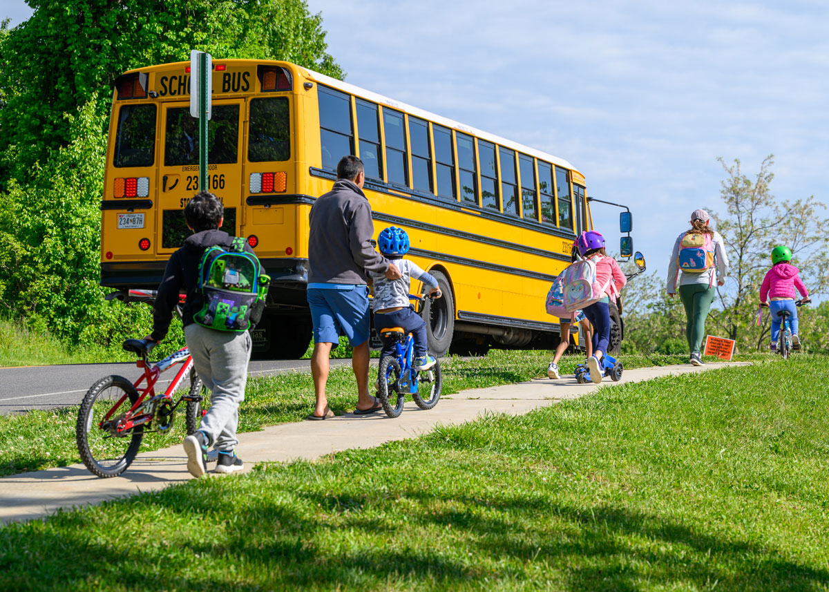 Image of childen and parents walking and cycling on a sidewalk with a school bus positioned on the adjacent street
